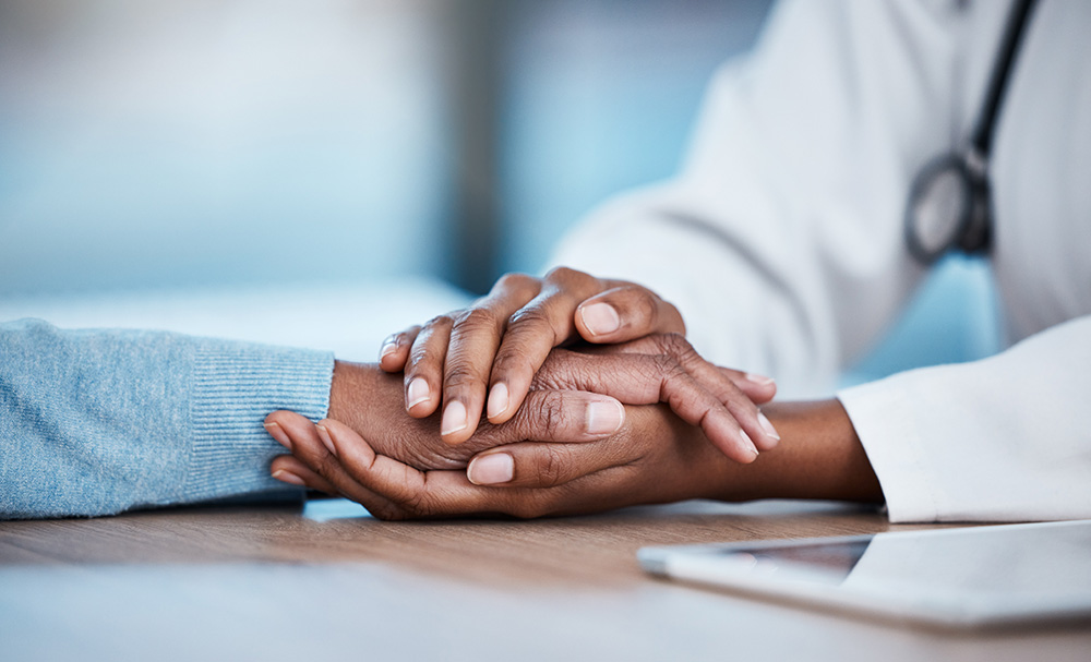 A closeup shows a doctor’s hands holding a patient’s across a desk.