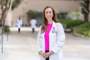 Dr. Danielle Sterrenberg stands outside in a pink shirt and a white medical coat and smiles.